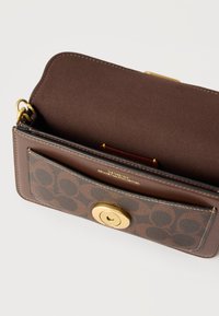 Καφέ τσάντα χιαστί από δέρμα με χαρακτηριστικό μονόγραμμα στην μπροστινή τσέπη, χρυσό κούμπωμα περιστροφής και εσωτερική επένδυση από σουέτ.