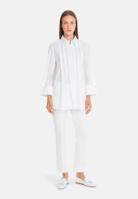 White, long-sleeve shirt with a stand-up collar, featuring vertical pleats and buttoned cuffs. Paired with white trousers and light shoes.
