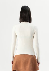 Femme avec des cheveux bruns à mi-longueur portant un haut blanc à manches longues côtelé et une jupe marron, se tenant de dos contre un fond uni.