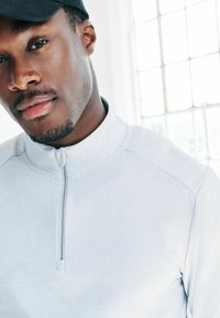 Tuvplāksnē redzams vīrietis ar melnu cepuri un gaiši pelēku blūzi ar rāvējslēdzēju, kas stāv blakus spožai logam ar kvadrātveida rūtīm.