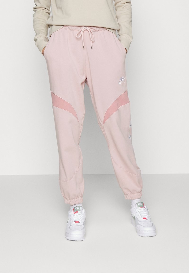 Osoba v světlých růžových Nike Air teplácích s elastickými manžetami, ruce v kapsách, doplněná bílými Nike Air teniskami a bílými ponožkami.