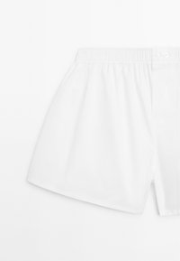 Bílé bavlněné boxerky s elastickým pasem a dvojitou knoflíkovou légu, ležící na bílém pozadí.