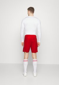 Witte lange mouwen sporttop, rode shorts met een losse pasvorm, witte sokken met rode strepen, en witte sportschoenen.