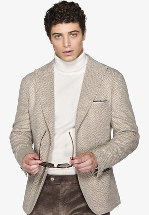 Blazer beige a spina di pesce con rever a punta, indossato sopra un dolcevita bianco. Il modello tiene occhiali marroni. Pantaloni in velluto a coste marroni.