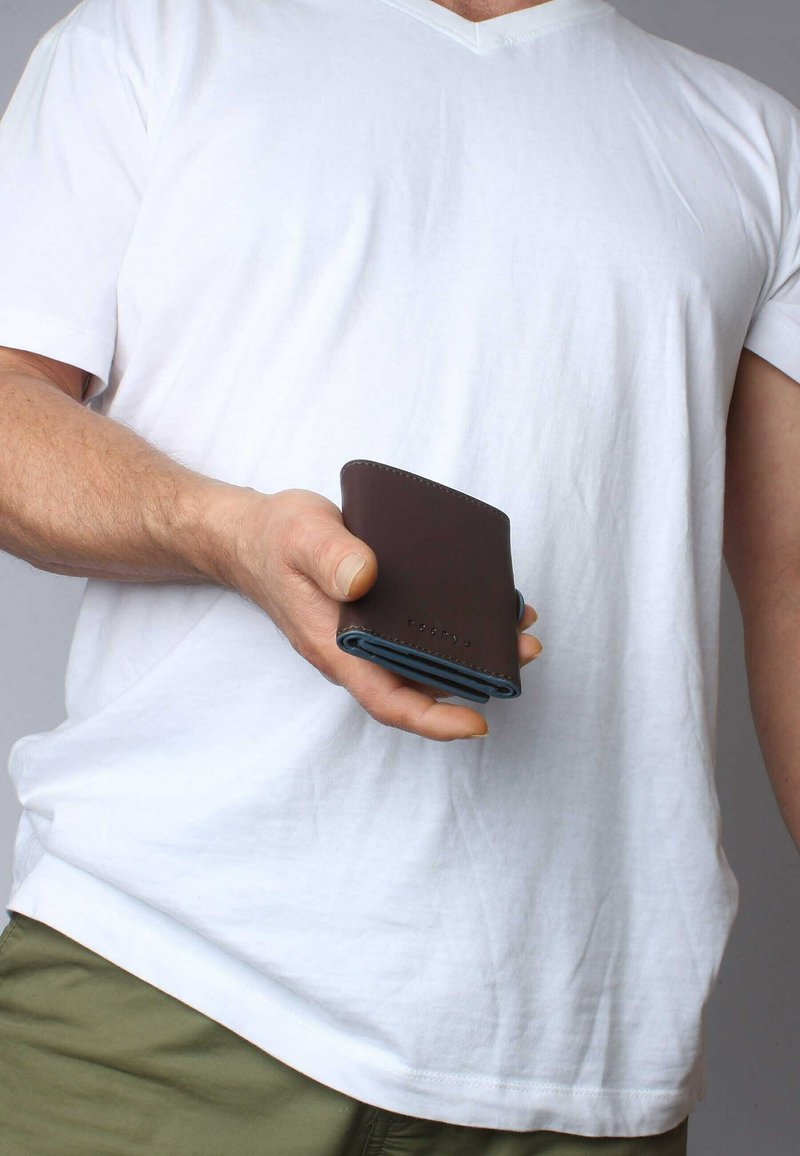 Portefeuille en cuir marron avec des accents bleus, au design compact et aux bords cousus, tenu dans une main. Il présente une texture lisse et un style minimaliste.