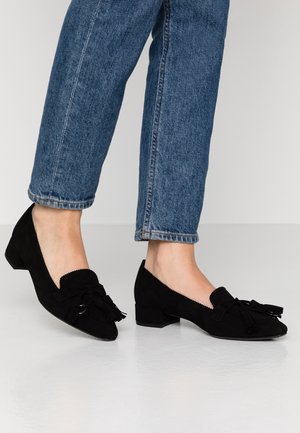 Person wearing blue denim jeans and black suede low-heeled loafers with tassel details standing on a light floor.