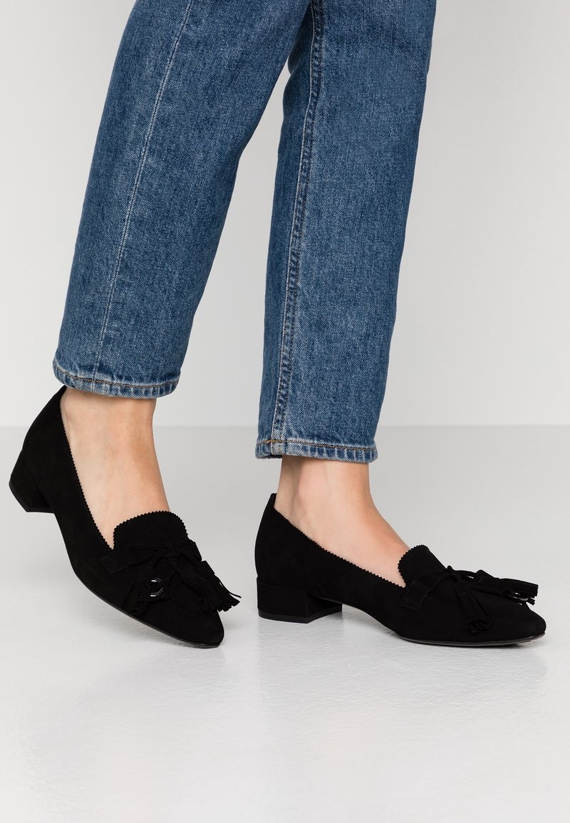 Personne portant un jean en denim bleu et des mocassins en daim noir à talon bas avec des détails à pompons, debout sur un sol clair.