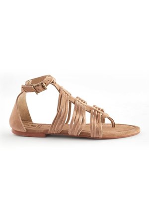 Sandale en cuir suédé nude avec une semelle plate, présentant des détails en frange sur le dessus, une sangle de cheville avec boucle et un design ouvertes à l'avant.