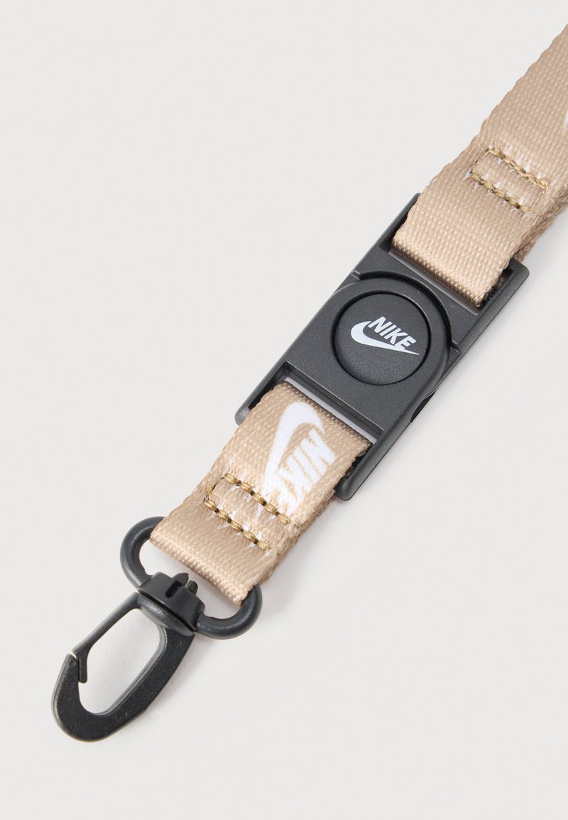 Lanyard beige de Nike con clip de plástico negro y gancho estilo mosquetón, con logo de Nike en la tela y botón circular.