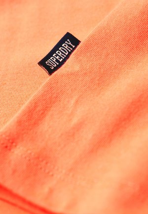 Tissu corail orange au toucher doux, avec une étiquette bleu foncé portant le mot "SUPERDRY" en lettres majuscules blanches.