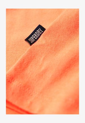 Tejido de color naranja coral con una textura suave, que presenta una etiqueta de color azul oscuro con el texto "SUPERDRY" en letras mayúsculas blancas.
