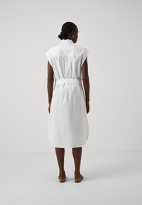 Abito bianco senza maniche con colletto, vita aderente con fiocco, lunghezza al ginocchio, tessuto texture e silhouette fluida. Sandali visibili ai piedi.