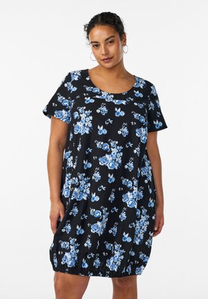 Frau trägt ein schwarzes, kurzärmeliges Kleid mit blauem und weißem Blumenmuster, steht vor einem schlichten weißen Hintergrund.