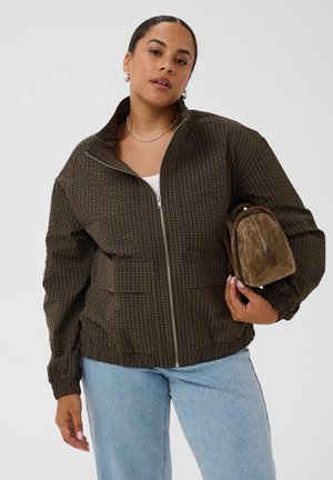 Femme portant une veste marron à carreaux par-dessus un haut blanc et un jean bleu clair, tenant un petit sac en daim marron sur un fond uni.