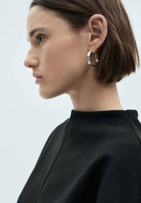 Silver hoop earrings with a polished finish; model wearing a black high-collared top. Background is neutral and minimalistic.