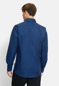 Mann mit kurzen braunen Haaren, der ein enganliegendes, langärmeliges blaues Hemd und schwarze Hosen trägt, von hinten vor einem schlichten Hintergrund zu sehen.