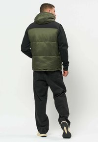 Un homme qui s'éloigne, portant une veste matelassée à capuche verte par-dessus un sweat noir et un jean noir ample avec des baskets à semelle beige.