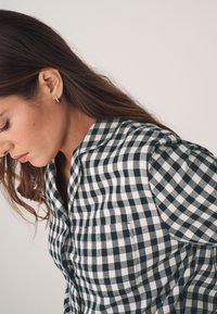 Kockás ing sötétzöld és fehér színben, kihajtós gallérral, gombos elülső résszel és zsebből a mellkason. Könnyű anyagból készült, puha tapintással.