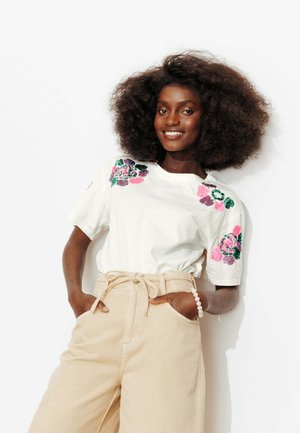 Femme souriante aux cheveux bouclés et volumineux portant un t-shirt blanc brodé de fleurs et un pantalon beige taille haute avec les mains dans les poches.
