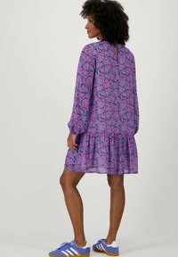 Paarse bloemenjurk met lange mouwen, gelaagde zoom en ronde halslijn met een sleutelgatdetail aan de achterkant. Gecombineerd met blauwe Sportschoenen.