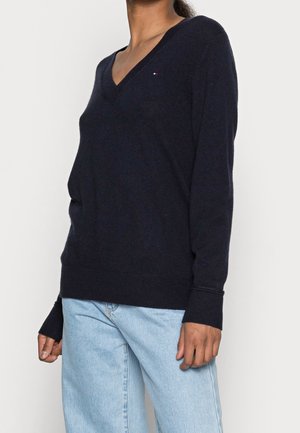 Maglione a maglia blu scuro con scollo a V, polsini e orlo a coste. Presenta un piccolo dettaglio con logo sul petto. Indossato sopra jeans azzurri chiari.