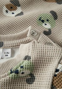 Tessuto beige con trama e motivi animali colorati, con bottoni a pressione in metallo sullo scollo. Etichetta con la taglia visibile.