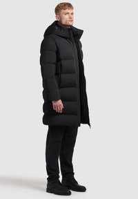 Black padded long coat with a hood, zipper closure, and side pockets. Smooth texture, featuring large quilted segments. Black denim pants and boots.