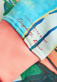 Met de hand beschilderde trui met een blauw en beige ontwerp, met een geribbelde roze boord. Geborduurde tekst in het rood met de woorden "Elizabeth Bowe".