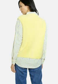 Geltonas megztinis be rankovių su rausvu tekstūru, dėvimas virš šviesiai mėlynos ir baltos juostuotos marškinės, derinamas su tamsiai mėlynomis džinsinėmis kelnių.