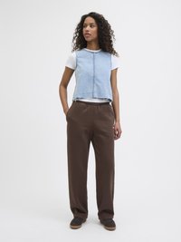 Lyseblå denim cropped vest over en hvit T-skjorte, paret med brune joggebukser. Modellen står med hendene i lommene og har på seg mørke sko.