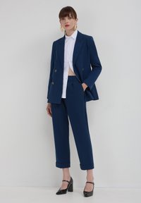 Abito blu navy su misura con giacca doppiopetto, camicia bianca accorciata sotto e scarpe nere con tacco alto. Tessuto liscio e taglio moderno.
