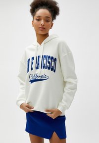 Biała bluza z kapturem z niebieskim nadrukiem "SAN FRANCISCO California", zestawiona z niebieską mini spódnicą o skośnym cięciu. Miękki, casualowy materiał.