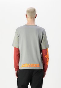 T-shirt in cotone grigio con logo e grafiche arancioni sul retro, con maniche lunghe rosse e strisce gialle sulle spalle.