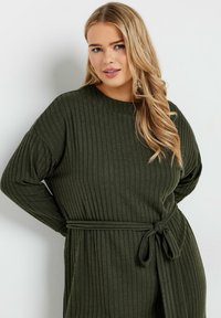 Robe tricotée côtelée vert foncé avec des manches longues et une ceinture nouée à la taille. Le tissu semble doux et texturé.