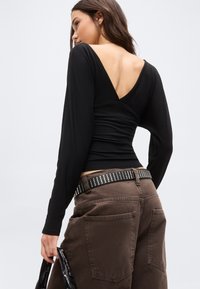 Frau trägt ein schwarzes Langarmshirt mit tiefem V-Rücken, braune Hosen mit großen Taschen und einen genoppten schwarzen Gürtel, hält eine schwarze Tasche.