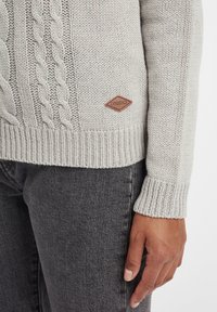 Light gray knitted sweater with cable knit pattern, ribbed cuffs, and a brown leather logo patch; worn over dark gray jeans.