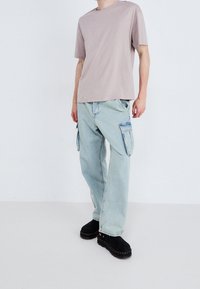 Jasno beżowy t-shirt z okrągłym dekoltem w połączeniu z jasnoniebieskimi spodniami cargo z bocznymi kieszeniami i luźnym krojem. Czarne buty wsuwane dopełniają stylizację.