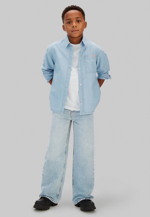 Junge steht mit hinter dem Rücken verschränkten Händen, trägt ein hellblaues Jeanshemd, weißes T-Shirt, weit geschnittene Jeans und schwarze Schuhe vor einem einfarbigen Hintergrund.