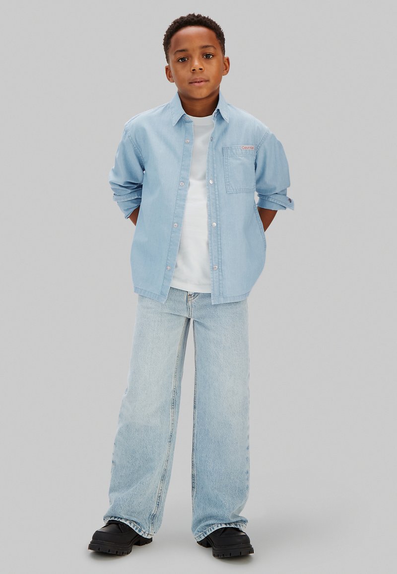 Garçon debout, les mains derrière le dos, portant une chemise en jean bleu clair, un t-shirt blanc, un jean à jambes larges, et des chaussures noires sur un fond uni.