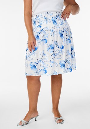 Femme portant une jupe blanche à motif floral bleu, un haut blanc et des sandales à talons ouvertes bleu clair, debout avec une main sur la hanche.
