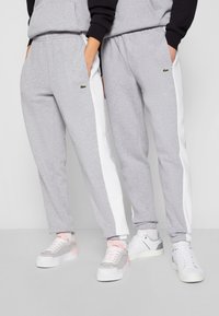 Grijze joggingsbroek met witte zijstrepen, voorzien van zakken en een klein groen logo. Gepaard met lichte sneakers, die verschillende texturen laten zien.