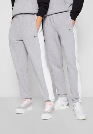 Pantalons de survêtement gris avec des rayures blanches sur les côtés, dotés de poches et d'un petit logo vert. Assortis à des baskets claires, mettant en valeur des textures variées.