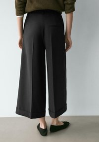 Schwarze, weit geschnittene Hosen mit poliertem Finish, mit umgeschlagenem Saum, einer flachen Gesäßtasche und glatter Textur, kombiniert mit dunkelgrünen Schuhen.