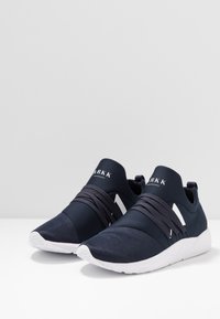 Baskets de sport bleu marine avec un design texturé, dotées de sangles élastiques sur le dessus et d'une semelle en caoutchouc blanc, portant la marque "ARKK" sur la languette.