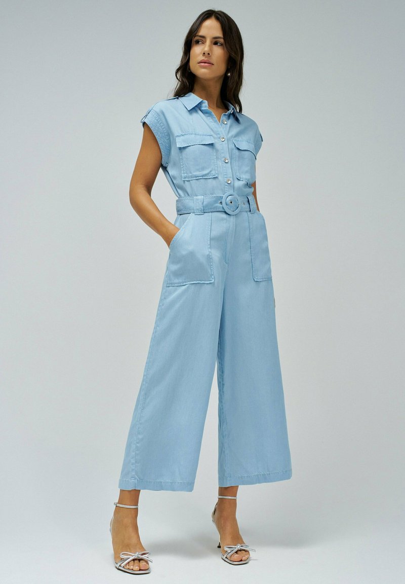 Jumper en denim bleu clair avec des manches courtes, un bouton à l'avant, deux poches poitrine, des jambes larges et une ceinture assortie. Sandales argentées à lanières.