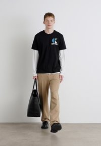 Sort T-shirt med logo, lagd over en hvid langærmet skjorte. Tan bukser, sorte sko og en stor sort tote-bag. Enkle farver, afslappet stil.