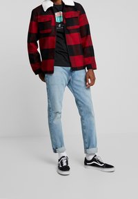 Henkilö, joka pukeutuu puna-mustaruudulliseen takkiin, jossa on valkoinen kaulus, musta kuvioitu t-paita, vaaleansiniset käännetyt farkut, valkoiset sukat ja mustat lenkkitossut.