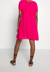 Pink short-sleeved dress with a loose, flowing design and gathered neckline, paired with black strappy sandals on bare feet.