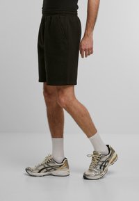 Homme marchant portant des shorts noirs, des chaussettes blanches montant jusqu'au mollet et des chaussures de course argent-beige sur un fond gris uni.