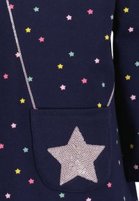 Tessuto blu navy con piccole stelle multicolori e una tasca scura con una grande stella in paillettes argentate e un motivo a catena sulle maniche.